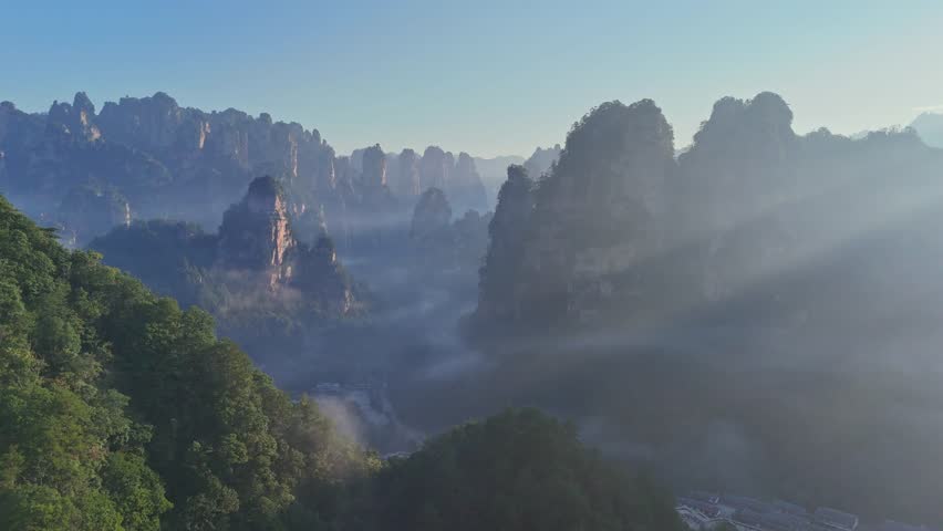 Stunning drone footage of the iconic sandstone pillars in Zhangjiajie, China, shrouded in morning fog with sun rays piercing through the valley. Perfect for travel documentaries and nature content.
