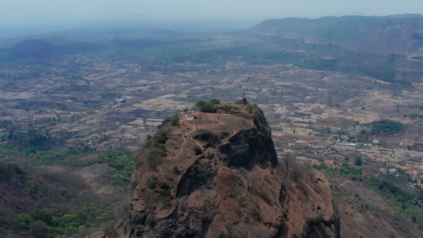 Slow drone orbit circles rugged Sarasgad Fort pinnacle topped by small temple amid sparse vegetation, overlooking vast arid plains and distant hazy hills from Pali village.
