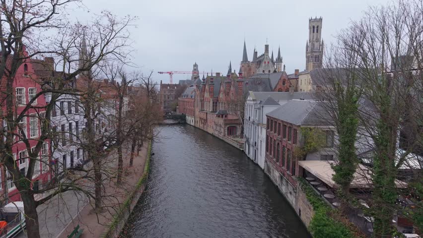 Drone reveal showing the historic canals and medieval architecture of Bruges, Belgium. Moody winter atmosphere with overcast sky and ancient brick buildings along the water.