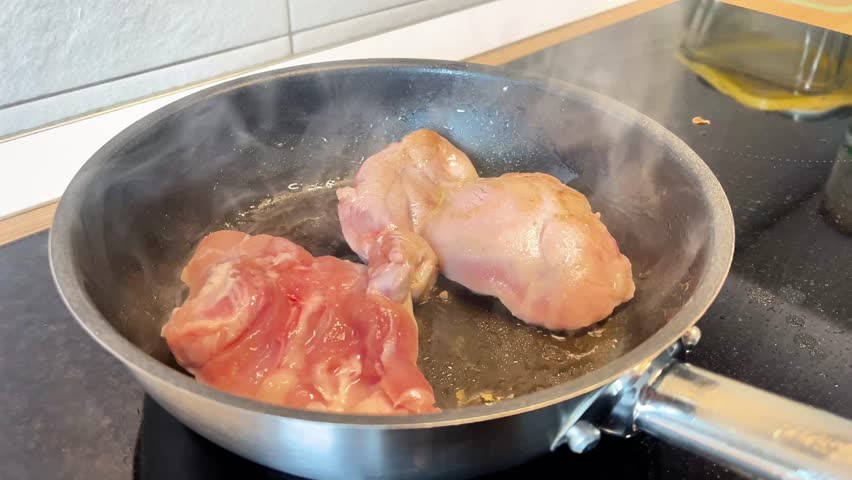 Two pieces of chicken are cooking in a pan on a stove. The pan is hot and the chicken is browning