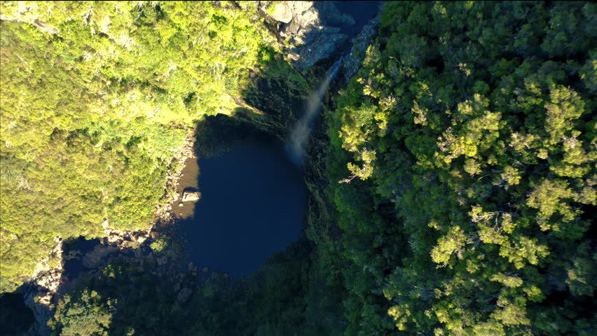 Aerial top down view of Lagoa do Vento waterfall in Madeira. Drone descending over hidden lagoon in deep shadows with bright sunlight hitting mountain cliffs in high contrast dramatic landscape.