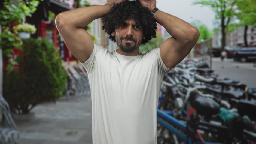 Man with curly hair smiling hands on head showing bare arms while standing near parked bicycles on a street in amsterdam; joy travel exploration.