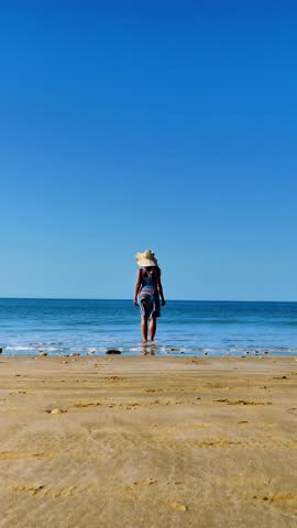 woman in dress and hat standing on the beach looking at the waves