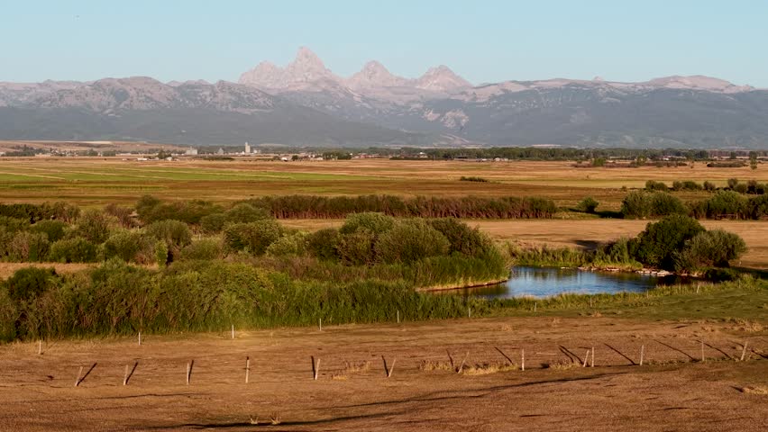 Aerial view of farmland with irrigation channels and grassy fields, small ponds reflecting the sky and Teton Mountain peaks in the background.