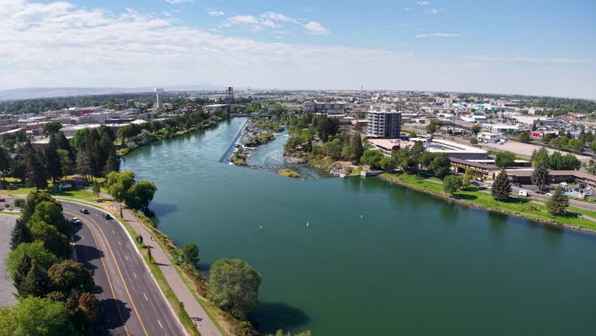 Aerial view of a wide river with a road along its bank and a distant city skyline under a clear blue sky.