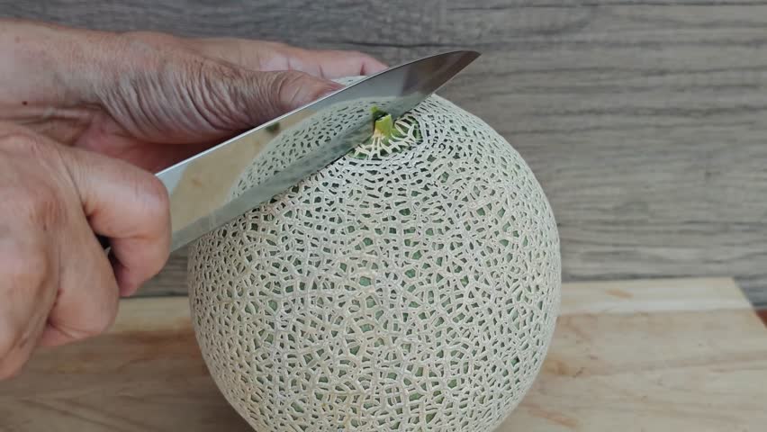 Close-up of a hand slicing a cantaloupe melon in half on a wooden cutting board.