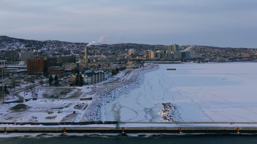 High aerial perspective of the Duluth city skyline and shoreline overlooking a completely frozen Lake Superior with prominent ice cracks at sunset.