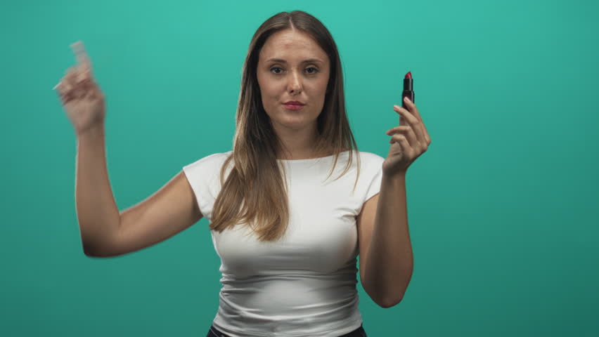 Woman brunette holding red lipstick and finger to temple in studio with teal backdrop wearing white t shirt, smiling with closed eyes; playful self care.