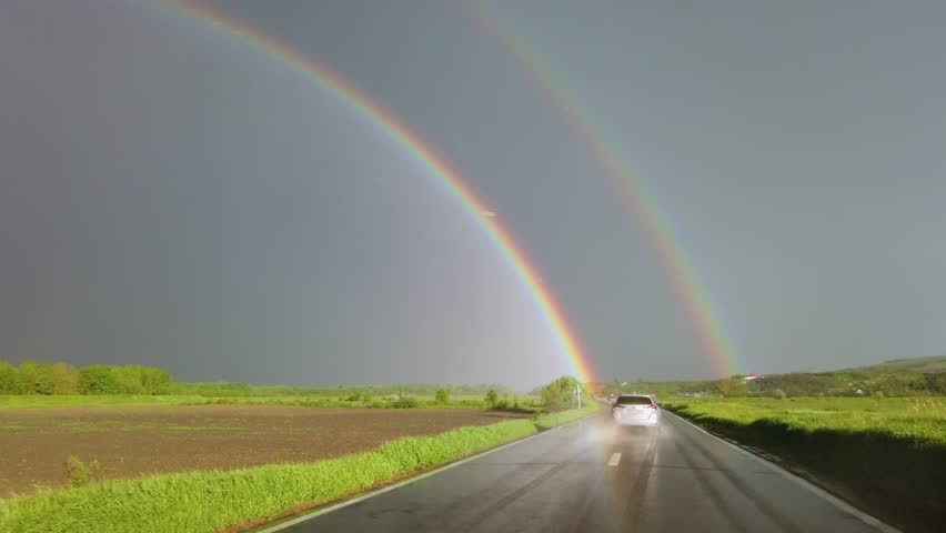 Double rainbow over wet asphalt road after spring rain with car driving towards horizon