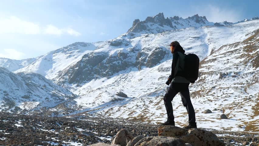 A male photographer is high in the mountains in winter, taking photos of snow-capped peaks. A guy with a camera and a backpack
