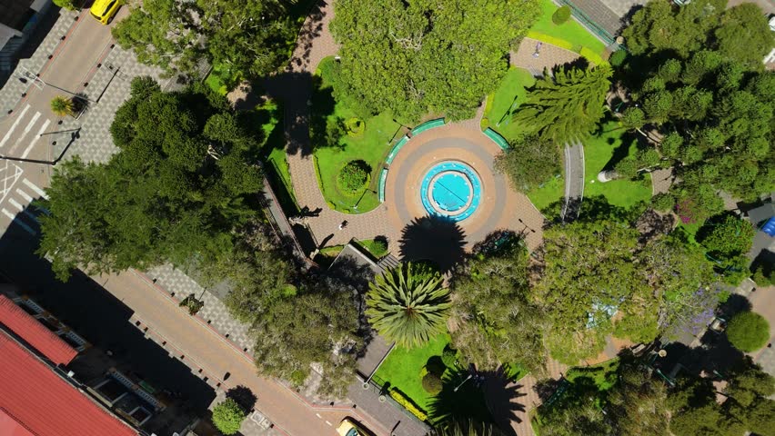 Top down drone shot rises above gardens and fountain of Palomino Flores Central Park revealing the surrounding city streets and architecture of Banos de Agua Santa, Ecuador