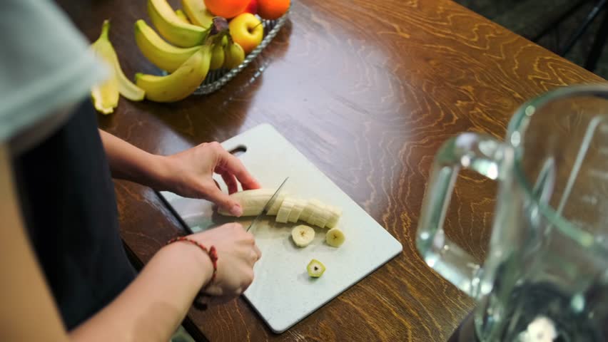 Female hands slice a ripe sweet banana into rounds on a cutting board, preparing a healthy fruit smoothie. On the wooden table is a plate with juicy oranges and fragrant apples.