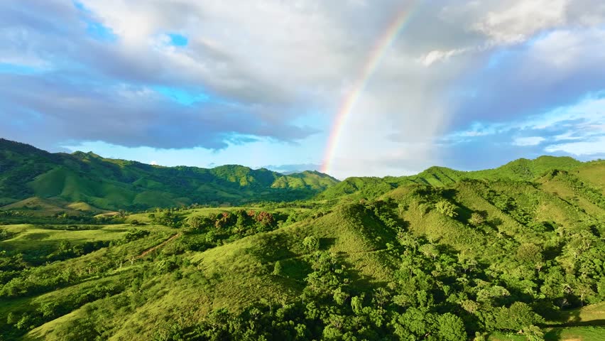 Mountain rainbow landscape under cloudy rainy sky. A natural, outdoor travel backdrop. Beautiful scenery with forests, hills, and meadows in the evening light. A ridge with a high peak in the distance.