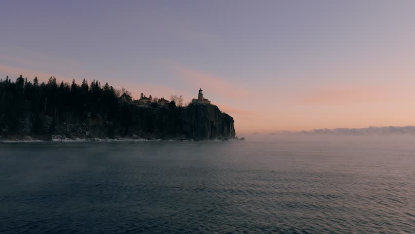 A serene dawn aerial of Split Rock Lighthouse silhouetted against a purple and pink winter sky over Lake Superior.