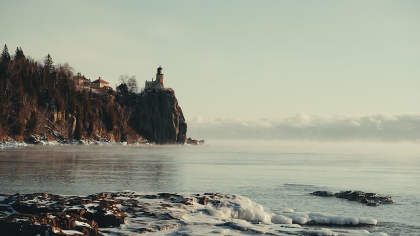 Split Rock Lighthouse stands on a rugged cliff over Lake Superior as winter sea smoke rises from the water at sunrise.