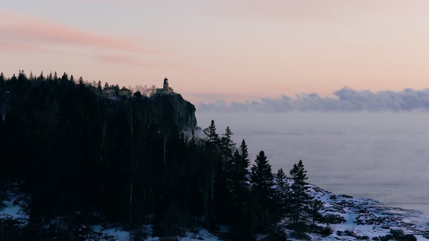 Aerial view looking through pine trees toward Split Rock Lighthouse as it stands guard over a misty winter lake.