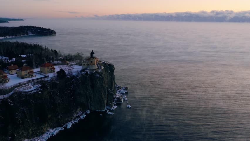 High-angle aerial view of Split Rock Lighthouse perched on an icy cliff overlooking a vast, misty Lake Superior at sunrise.