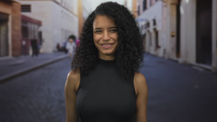 Woman with curly hair pointing fingers playfully on a city street during daylight, capturing a joyful expression, embodying urban vibrancy and lively interaction.
