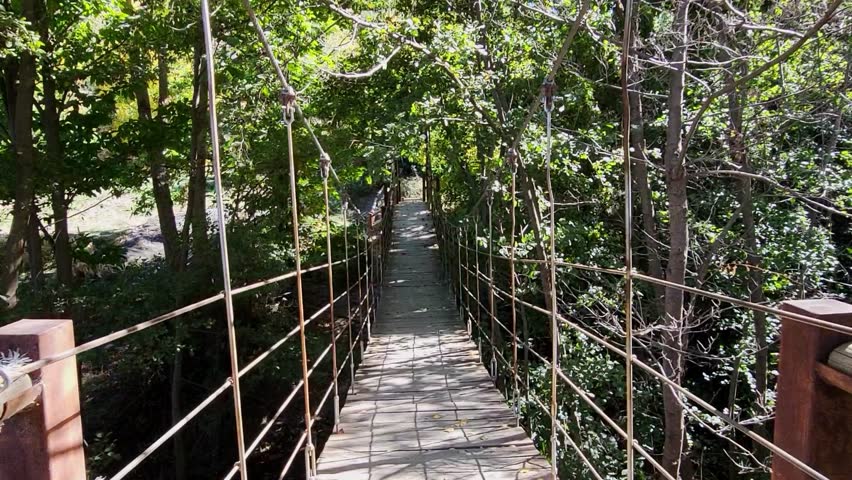 Walking across a suspension bridge over the river. Hanging walkway between trees. First-person view. Spain.