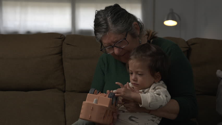 Short lifestyle clip of a Spanish grandmother sitting on a sofa with a half- Asian young child while the child spins the wheels of a toy truck during playtime at home