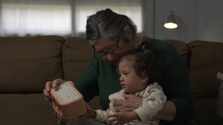 Close-up lifestyle shot of a grandmother playing with a toy truck as a young child laughs joyfully beside her on a sofa at home, capturing a warm multi-generational moment
