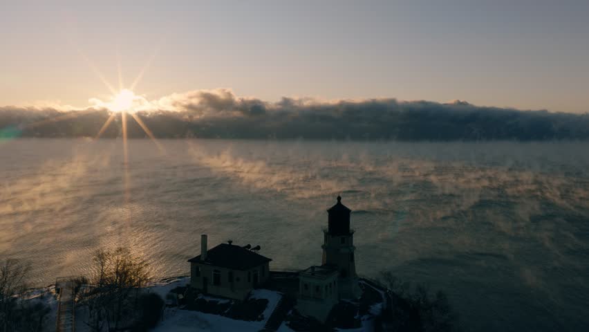 High-angle drone shot of the rising sun over a misty Lake Superior and the historic Split Rock Lighthouse in winter.
