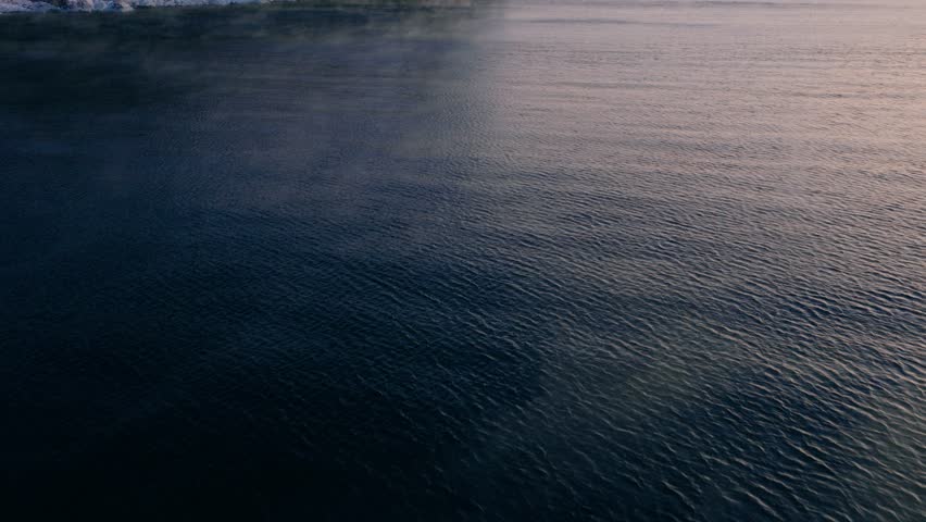 Low-angle aerial approach over the rippling water of Lake Superior, revealing Split Rock Lighthouse through thick morning sea smoke.