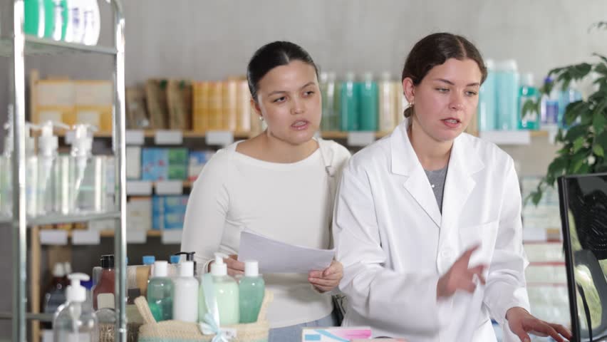 Attentive young female pharmacist working on computer while Asian woman asking for prescribed medicaments chemistry 