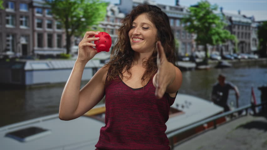 Young woman holding a red piggy bank and extending her right hand for a handshake on a street by a canal and building facades in amsterdam; saving optimism.