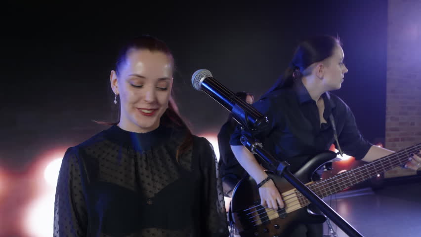 Closeup of caucasian young woman in black t shirt playing keyboard and electric bass guitar on stage indoors with blue lighting, performing energetic rock music.