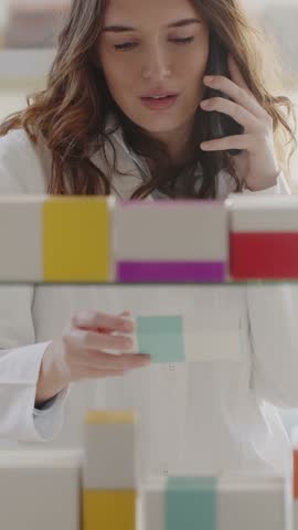 Female pharmacist selecting the correct medicine from pharmacy shelves while talking on a smartphone, providing professional, attentive, and trustworthy customer service.
