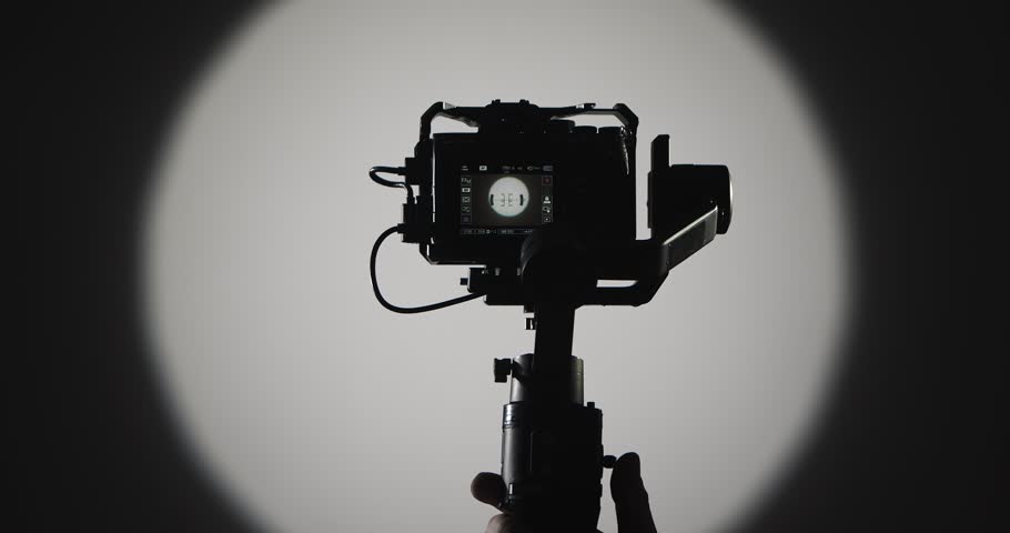 The operator stabilizes the camera mounted on a motorized stabilizer with a suspension, filmed against a bright round light on a dark background, symbolizing film production.