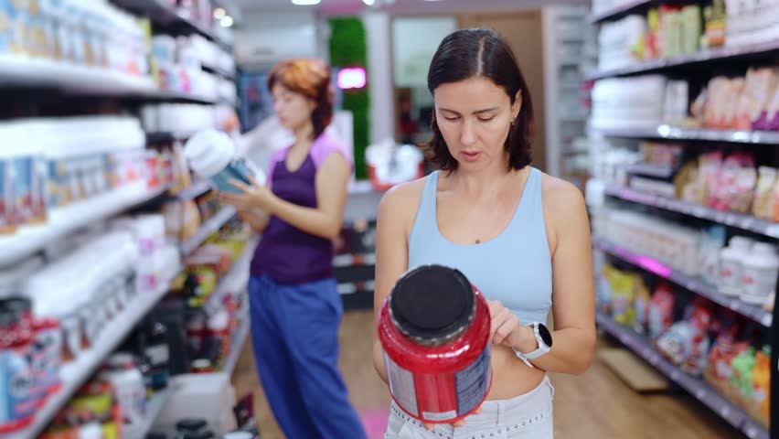 Thoughtful female customer reviewing jars of protein standing among shelves in sports food shop