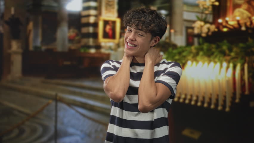 Teenager covers face with hands beside lit votive candles in a historic church building wearing striped shirt and curly hair showing nervous smile; surprise.
