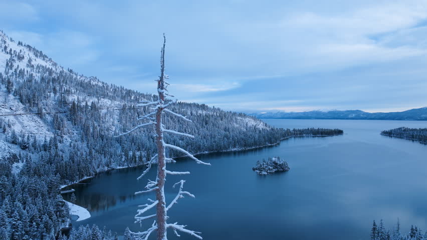 Aerial drone footage of Lake Tahoe surrounded by snow covered forest and winter shoreline. Calm blue water contrasts with frozen landscape,showcasing serene alpine nature and seasonal mountain scenery