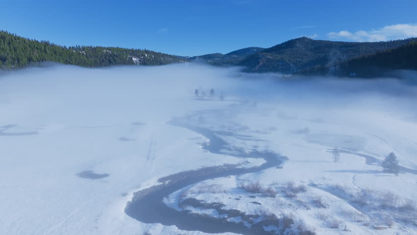 Aerial footage of morning fog filling a frozen valley with a winding stream, snowy fields, pine trees and mountain ridges near Palisades Tahoe, California, USA. Peaceful alpine winter scene.