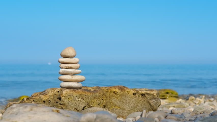 Balanced stone pyramid on a seashore. Serene stack of smooth pebbles balancing on a larger rock on a quiet beach, symbolizing harmony, stability, and tranquility with the calm blue ocean and clear sky