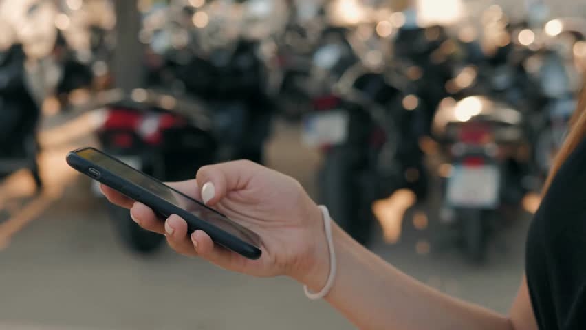 Focus on woman interacting with phone in outdoor setting, with rows of parked scooters in background at outdoor parking space. The sun lights up the scene.