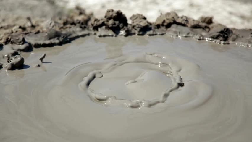 A small mud volcano actively bubbles and erupts in a pool of gray mud. Close-up view of the dynamic landscape during daytime at an active site.