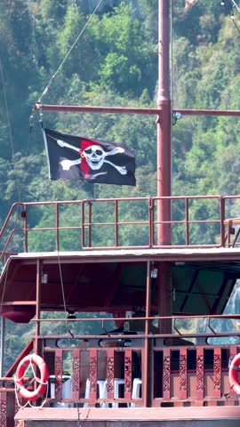 A black Jolly Roger flag waves from a wooden ship mast against lush tropical cliffs