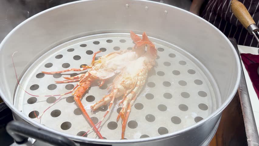 A spiny lobster cooks inside a metal steamer basket with rising steam at street market