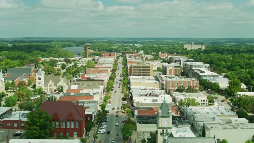 Aerial view captures downtown Lawrence, Kansas, showing city buildings interspersed with green trees under bright daylight, highlighting urban life.