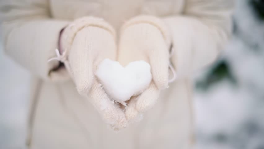 A close up view of female hands wearing white knitted gloves holding snow in shape of a heart during winter season, perfect for depicting love and warmth during cold weather with a soft and blurred background capturing the gentle falling snow