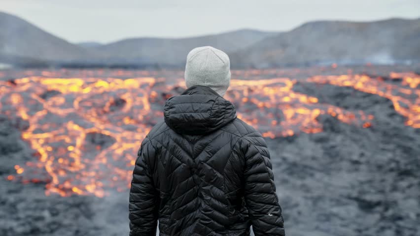 A person with warm winter clothes observes the fiery lava flowing from Fagradalsfjall Volcano across the landscape in Iceland. Amazing nature background.