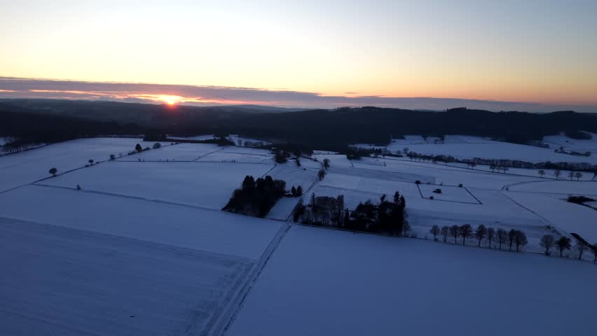 Dramatic sunset behind forest hills on snowy winter evening in USA. Aerial lateral wide shot. Blue hour scene on snow-covered farmland fields. Quiet and peaceful rural landscape in America.
