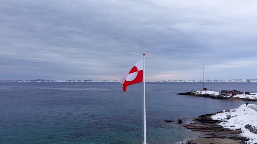 The red and white flag of Greenland (Erfalasorput) flutters in the wind against a backdrop of icy blue waters and snow-capped mountains in a serene Arctic landscape.