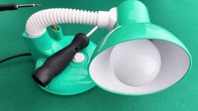 A green desk lamp with a flexible neck sits on a solid surface, its base holding a screwdriver. The lamp features a round button and bulb, highlighting a simple workspace repair setup - Powered by Shutterstock - Get 15% off with code: PIKWIZARD15