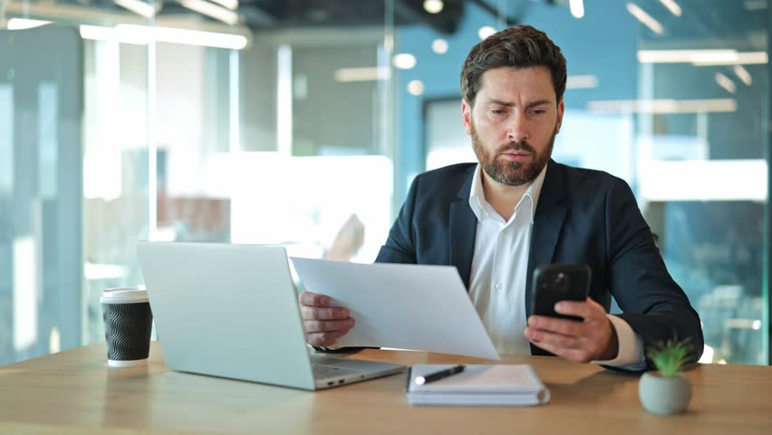 Professional man intently reviews documents, comparing information on smartphone and laptop. Demonstrates serious focus on business tasks and modern workplace efficiency, feeling concentrated.