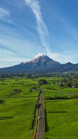 Vertical aerial hyperlapse of cars driving along a rural road with Mount Taranaki in the background. Snow-capped peak under blue sky on a sunny summer day in New Zealand, filmed along Kent Road.