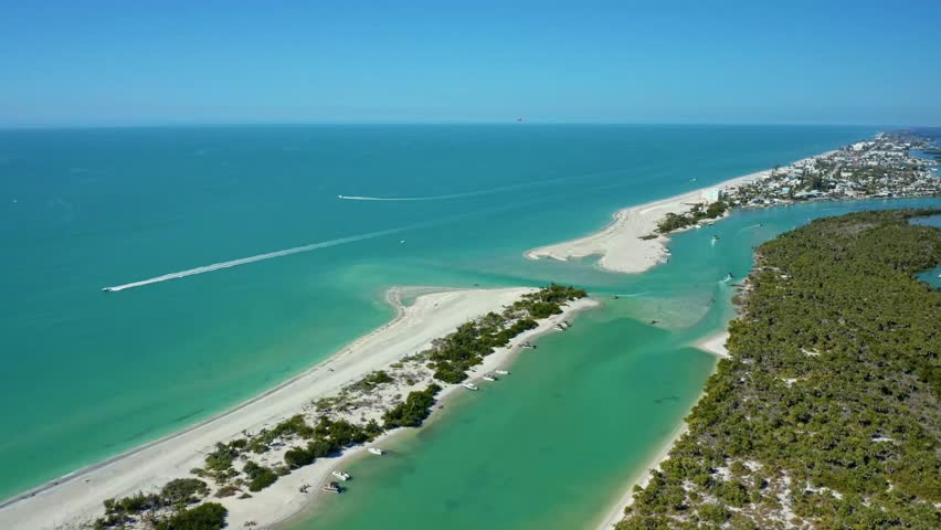 Coastal aerial view of a Florida shoreline featuring clear turquoise waters, white sandy beaches, several boats anchored near the shore, and urban development in the background under a clear sky.
